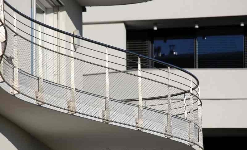 Geschwungenes Balkongeländer aus Edelstahl mit Drahtseilnetz-Füllung an einem weißen Neubau